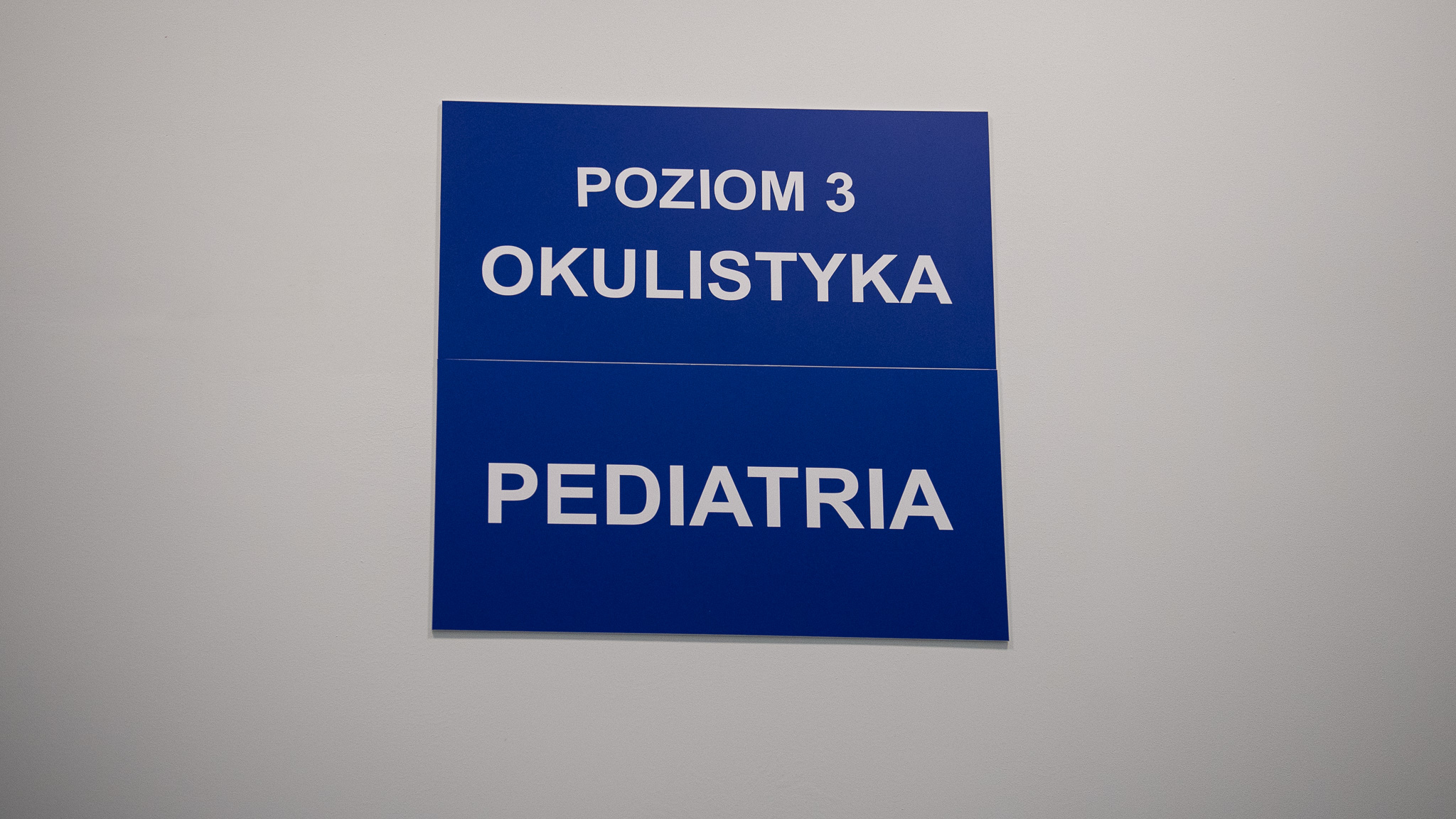 Dwie granatowe tablice informacyjne wewnątrz szpitala w Legionowie. Na pierwszej u g ory napis Poziom 3 Okulistyka, na drugiej poniżej napis Pediatria.