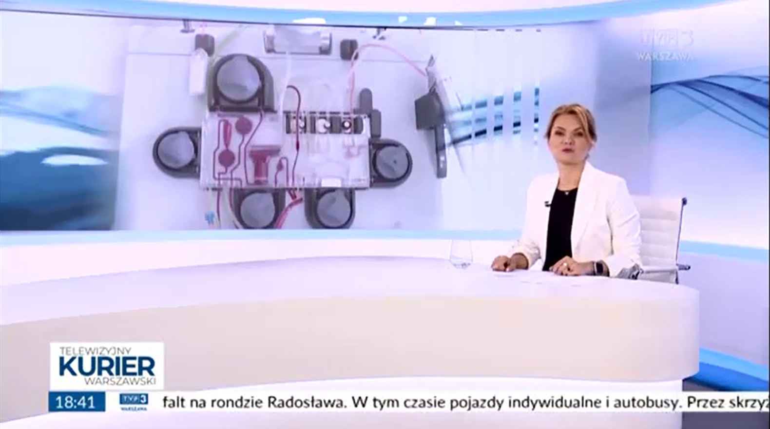 Zrzut ekranu programu telewizyjnego Telewizyjny Kurier Warszawski. Na pierwszym planie prezenterka siedzi przy białym stole w studio i prezentuje wiadomości