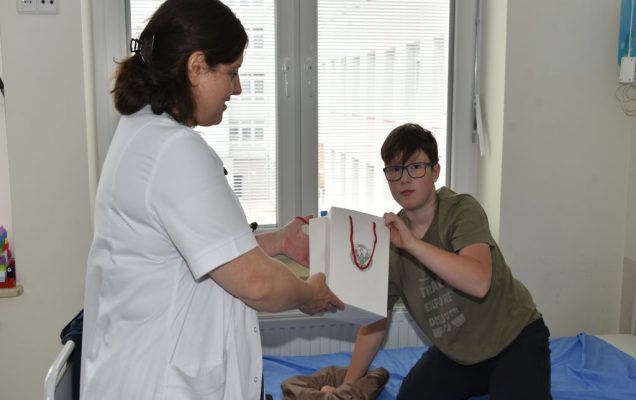 Dzień Dziecka w Klinice Pediatrii, Nefrologii i Alergologii Dziecięcej WIM-PIB, kilkunastoletni pacjent na sali szpitalnej siedzi na łóżku i odbiera prezent od pracowniczki kliniki.