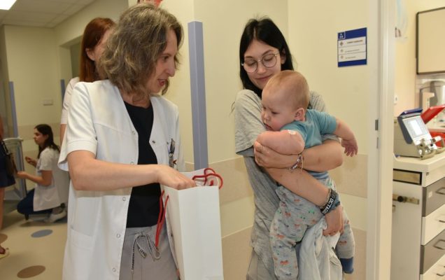 Dzień Dziecka w Klinice Pediatrii, Nefrologii i Alergologii Dziecięcej WIM-PIB, mama z niemowlakiem na rękach stoi uśmiechnięta na szpitalnym korytarzu i odbiera prezent od pracowniczki kliniki.