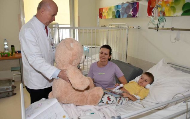 Kolorowe zdjęcie. Profesor Bolesław Kalicki kierownik Kliniki Pediatrycznej w sali Kliniki Pediatrycznej. Na łóżku leży mały chłopiec, obok siedzi jego mama. Profesor trzyma na łóżku olbrzymiego pluszowego misia którego da za chwilę chłopcu.