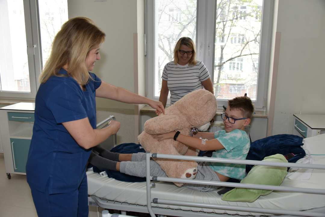 Kolorowe zdjęcie. Uśmiechnięta pracowniczka Kliniki Pediatrii wręcza uśmiechającemu się chłopcu leżącemu na łóżku szpitalnym dużego, pluszowego, beżowego misia. Za łóżkiem chłopca stoi usmiechnięta mama chłopca.