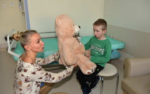 Kolorowe zdjęcie. Sala szpitalna. Małgorzata Złotkowska kucając wręcza małemu pacjentowi siedzącymi na taborecie dużego pluszowego beżowego misia.