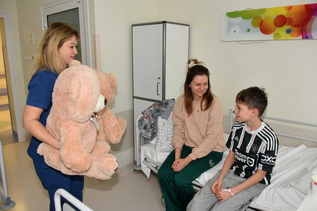 Kolorowe zdjęcie. Pacjent Kliniki Pediatrycznej z mamą siedzą uśmiechnięci na łóżku szpitalnym. Uśmiechnięta pracowniczka kliniki za chwilę wręczy chłopcu dużego pluszowego, beżowego misia.