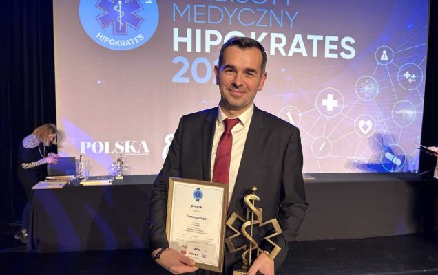 Tomasz Zając podczas Gali Hipokrates. W rękach trzyma dyplom oraz statuetkę Hipokrates 2024. Z tyłu scena i napis Plebiscyt Medyczny Hipokrates 2024