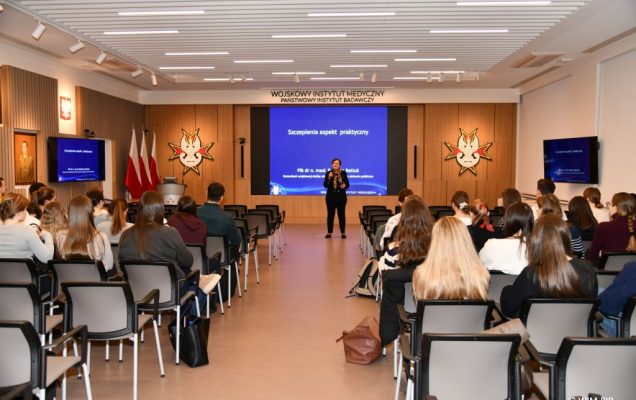 Widok z końca auli. Barbara Betiuk podczas szkolenia dla wojskowych lekarzy stażystów w Wojskowym Instytucie Medycznym stoi przed katedrą , w tle za nią wyświetlana z rzutnika prezentacja. Na prezentacji napis 