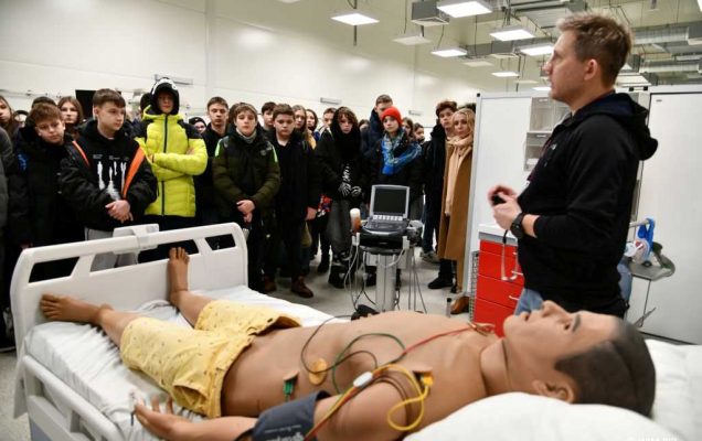 Pracownik Wim obok niego na łóżku leży fantom szkoleniowy podłączony do aparatury medycznej. Pracownik prowadzi szkolenie. Na przeciw pracownika grupa słuchających uczniów.