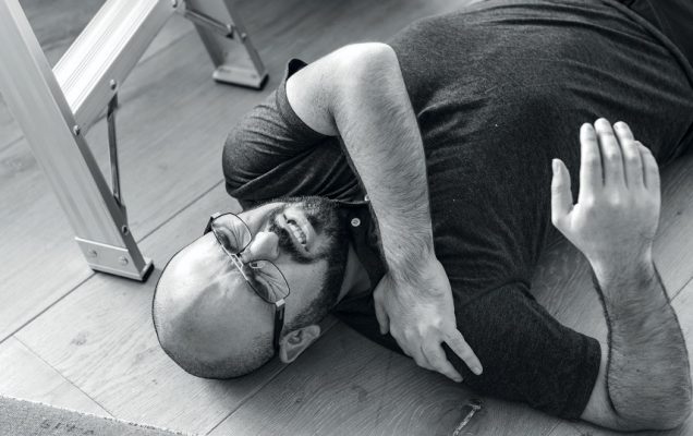 Zdjęcie czarno-białe. Leżący na podłodze w mieszkaniu mężczyzna trzymający się za bark.