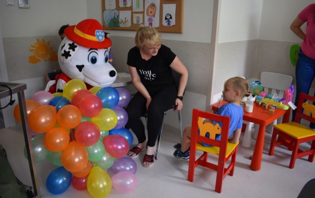 Sala pacjentów, maskotka Marshalla z balonami, obok siedzi mama i kilkuletni chłopiec.