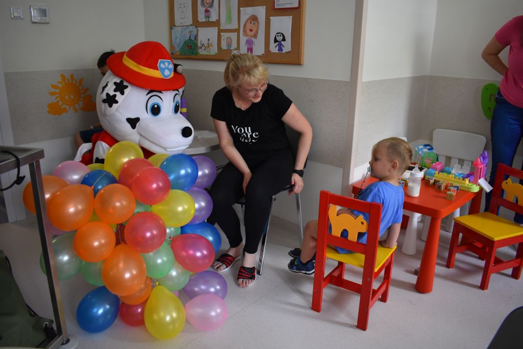 Sala pacjentów, maskotka Marshalla z balonami, obok siedzi mama i kilkuletni chłopiec.