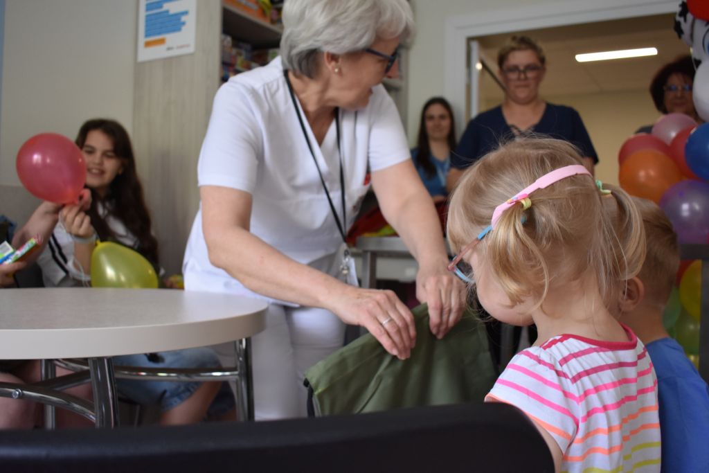 Pracownica szpitala i małą dziewczynka. Kobieta siedzi na krzesełku i wręcza prezenty.