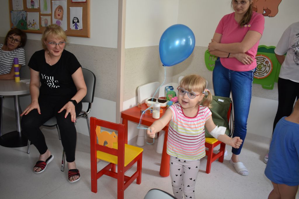 Kilkuletnia dziewczynka w sali szpitalnej stoi z niebieskim balonikiem w ręce.