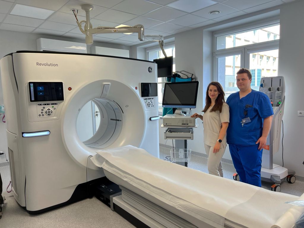 Dwóch pracowników służby zdrowia stoi obok tomografu komputerowego w sali szpitalnej wyposażonej w monitory i sprzęt medyczny.