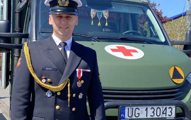 Żołnierz ubrany na galowo, za nim ambulans wojskowy.