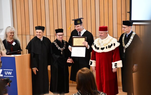 Wykładowcy uniwersyteccy ubrani w togi, obok nich student prezentujący dyplom.