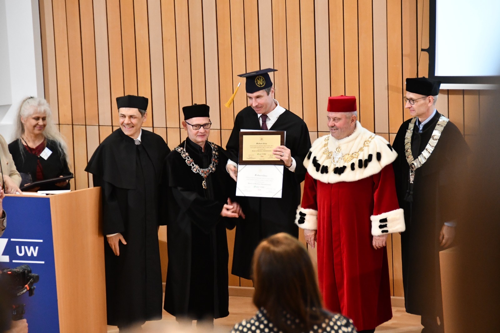 Wykładowcy uniwersyteccy ubrani w togi, obok nich student prezentujący dyplom.