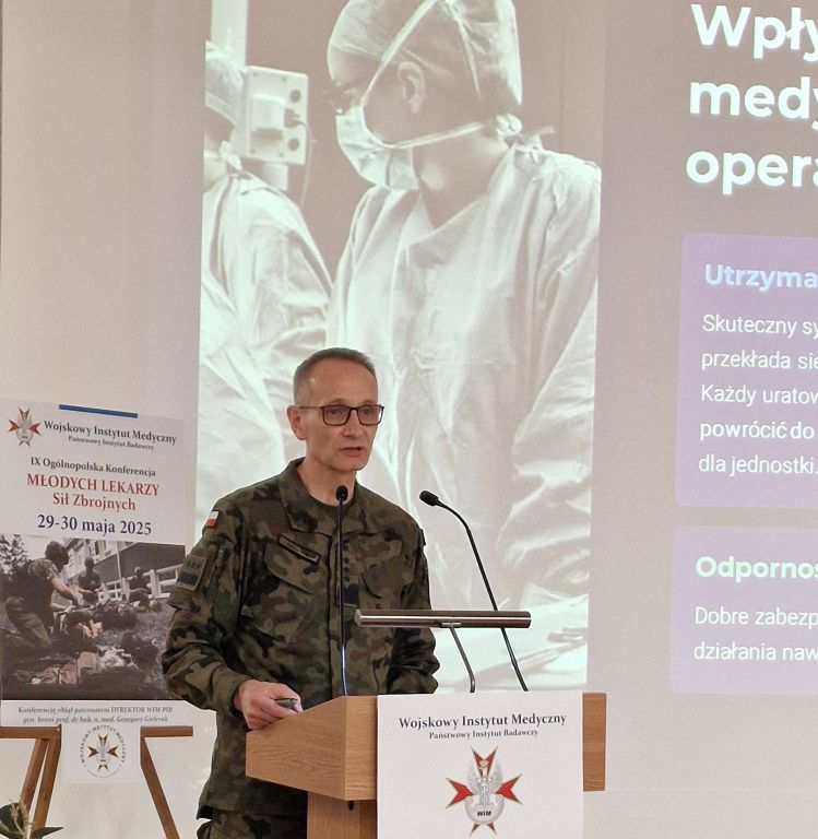 Dyrektor WIM-PIB w mundurze wojskowym przy mównicy. Za nim wyświetlony obraz z prezentacji lekarza w maseczce chirurgicznej.