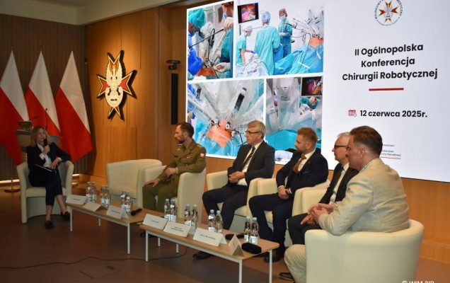 Debata konferencyjna. Prelegenci siedzący w fotelach, za nimi wyświetlony slaid reklamujący konferencję.