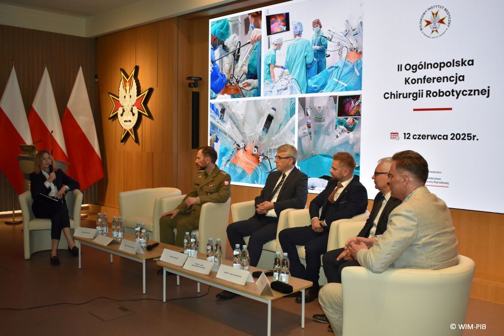 Debata konferencyjna. Prelegenci siedzący w fotelach, za nimi wyświetlony slaid reklamujący konferencję.