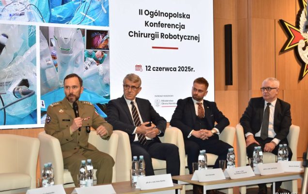 Debata konferencyjna. Prelegenci siedzący w fotelach, za nimi wyświetlony slaid reklamujący konferencję.