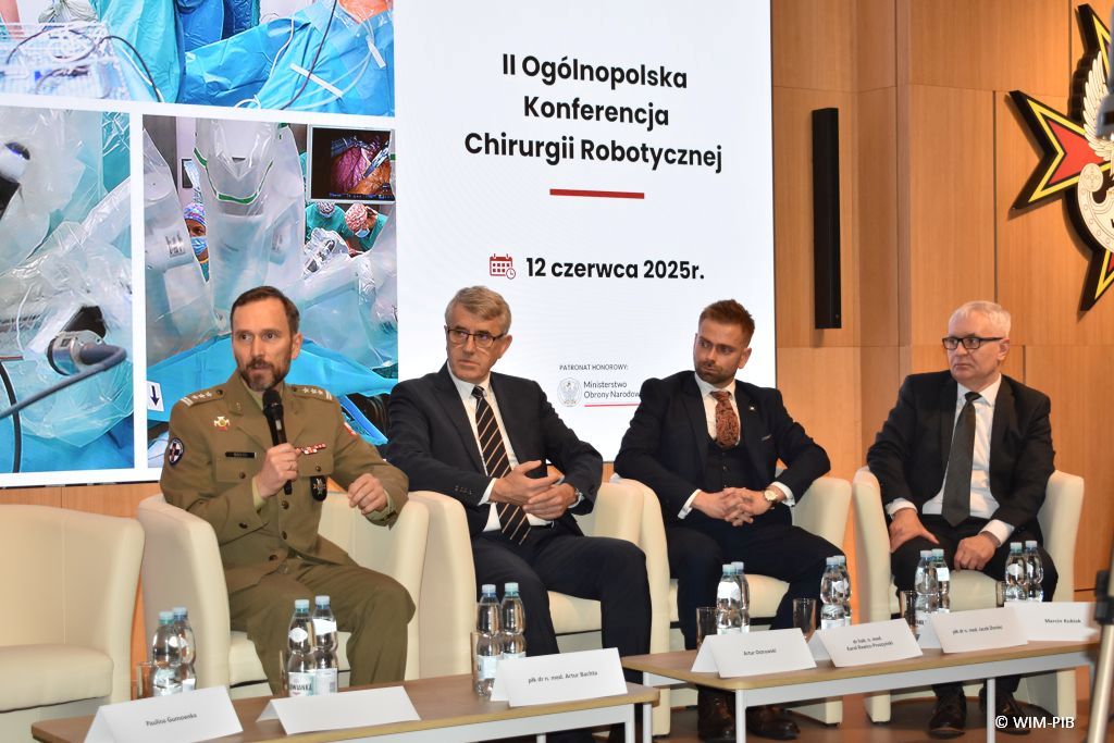 Debata konferencyjna. Prelegenci siedzący w fotelach, za nimi wyświetlony slaid reklamujący konferencję.