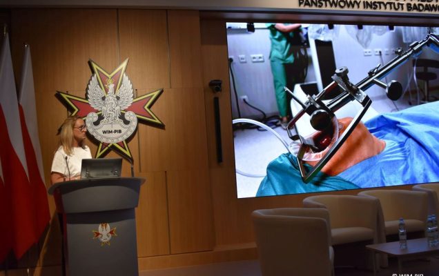 Dr n. med. Aldona Chloupek z mikrofonem przy mównicy podczas wykładu. Z jej lewej stronie ekran a na nim slaid z wykłądu.