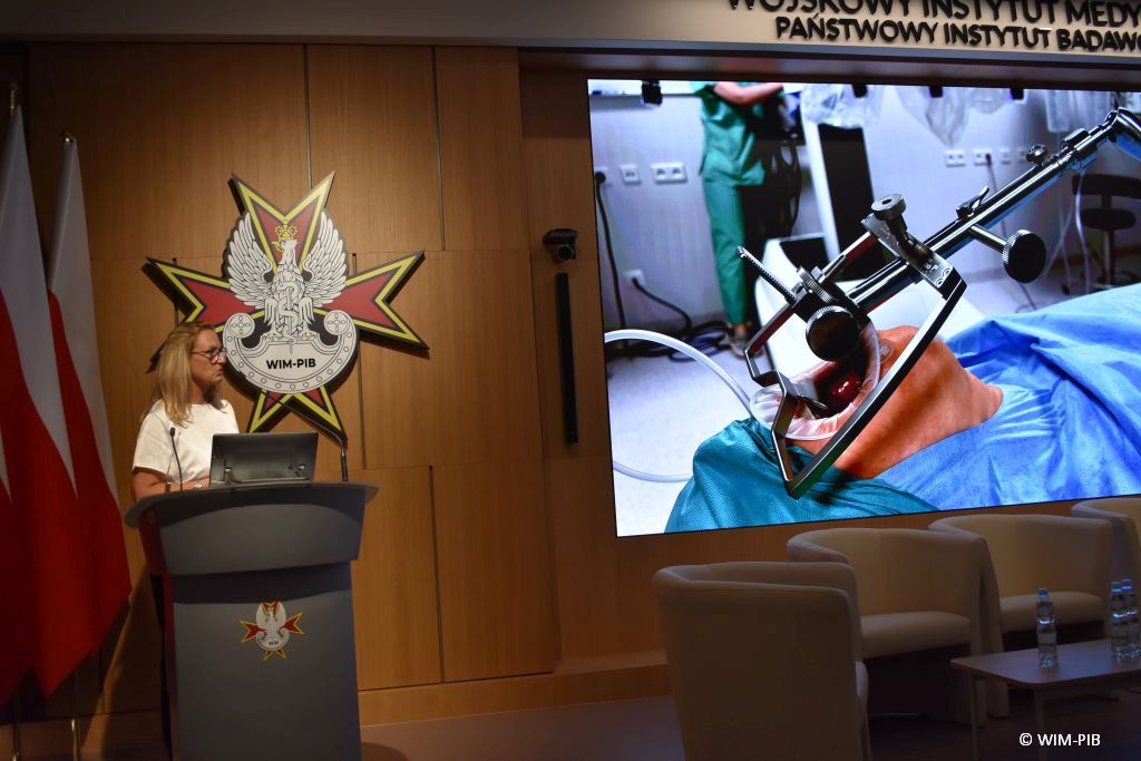 Dr n. med. Aldona Chloupek z mikrofonem przy mównicy podczas wykładu. Z jej lewej stronie ekran a na nim slaid z wykłądu.