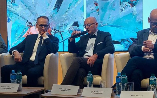 Debata konferencyjna. Prelegenci siedzący w fotelach, za nimi wyświetlony slaid reklamujący konferencję.