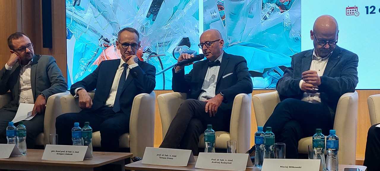 Debata konferencyjna. Prelegenci siedzący w fotelach, za nimi wyświetlony slaid reklamujący konferencję.