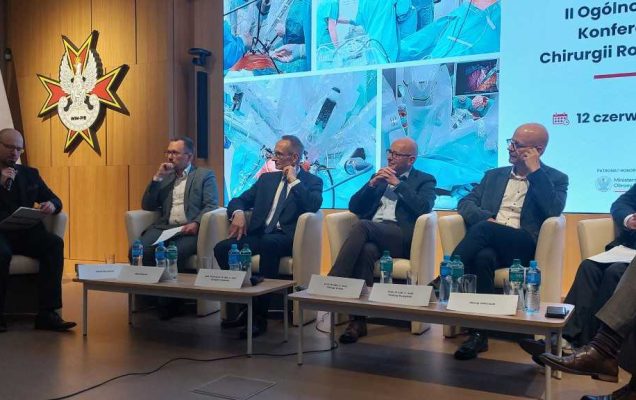 Debata konferencyjna. Prelegenci siedzący w fotelach, za nimi wyświetlony slaid reklamujący konferencję.