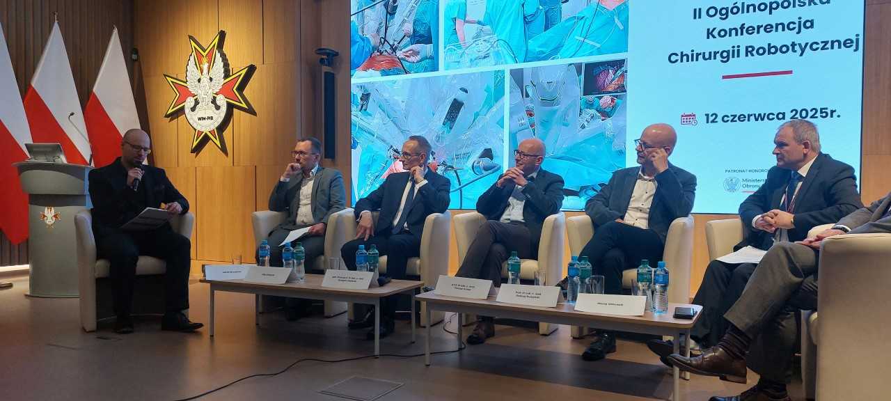Debata konferencyjna. Prelegenci siedzący w fotelach, za nimi wyświetlony slaid reklamujący konferencję.