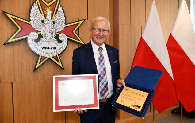 prof. dr hab. n. med. Wojciech Marczyński na auli WIM trzymający dyplom i nagrodę. Za nim logo WIM i flagi Polski.