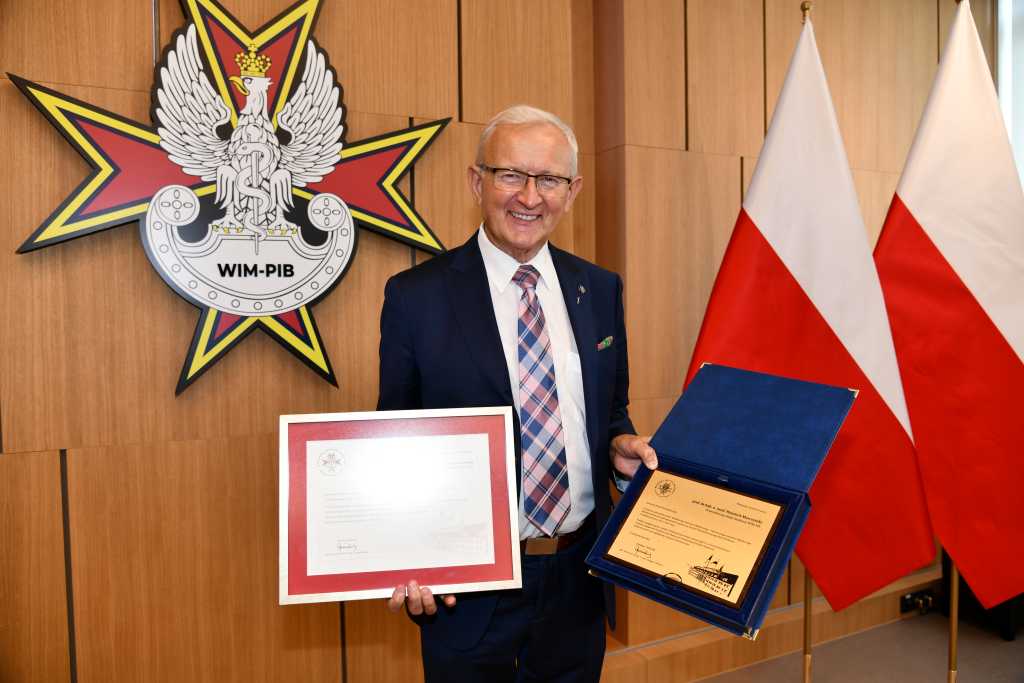 prof. dr hab. n. med. Wojciech Marczyński na auli WIM trzymający dyplom i nagrodę. Za nim logo WIM i flagi Polski.