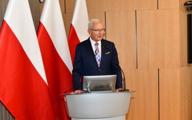 Prof. dr hab. n. med. Wojciech Marczyński na auli WIM przy mównicy, za nim flagi Polski.