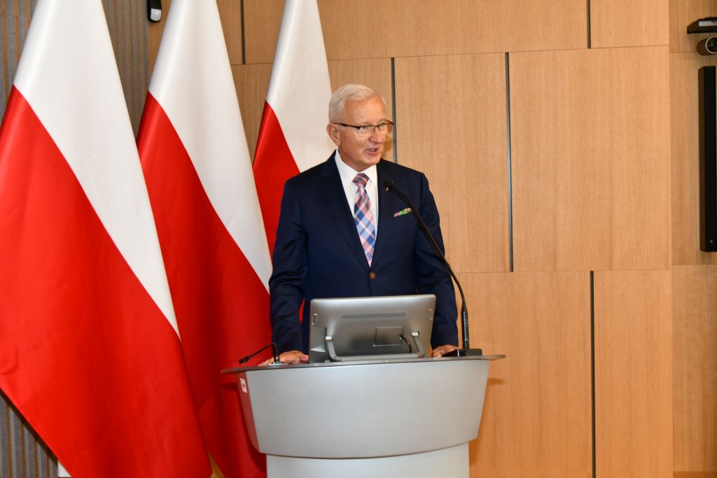 Prof. dr hab. n. med. Wojciech Marczyński na auli WIM przy mównicy, za nim flagi Polski.