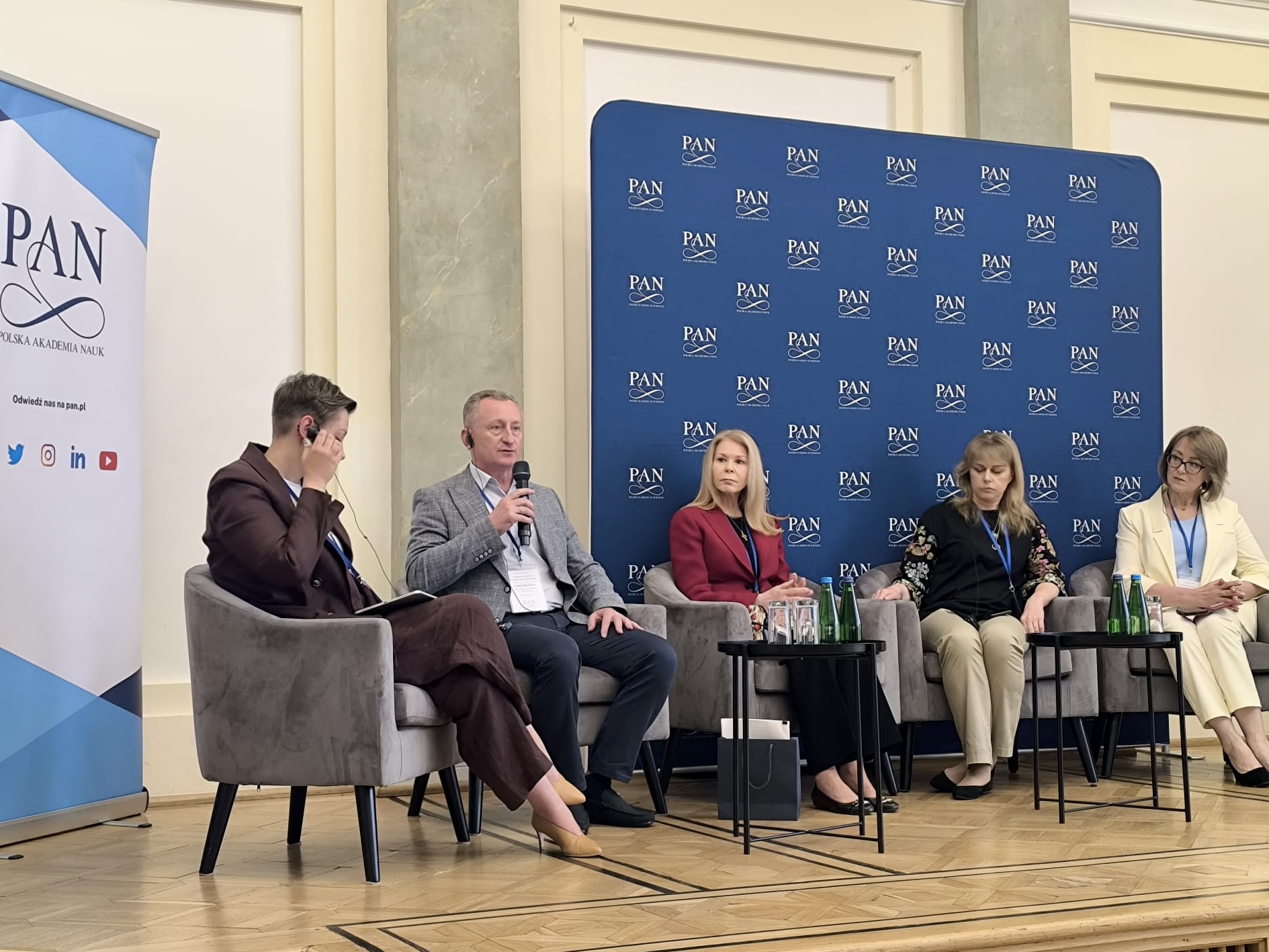 Panel dyskusyjny konferencji, siedzący prelegenci z animi tablica z logotypami PAN.