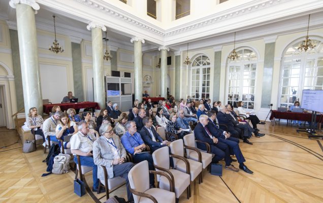 Uczestnicy konferencji siedzący w zabytkowej sali z kolumnami.