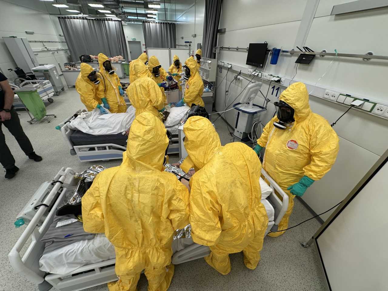 Medycy w żółtych kombinezonach i maskach przeciwgazowych podczas kursy CBRN w żółtych kombinezonach na sali szpitalnej.