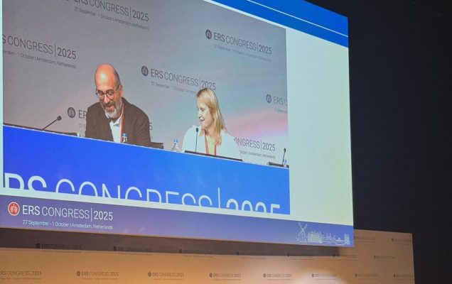 Prelegenci podczas konferencji. Nad nimi olbrzymi ekran wyświetlający ich obraz.