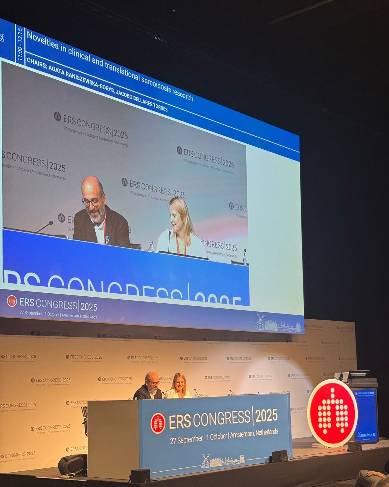 Prelegenci podczas konferencji. Nad nimi olbrzymi ekran wyświetlający ich obraz.
