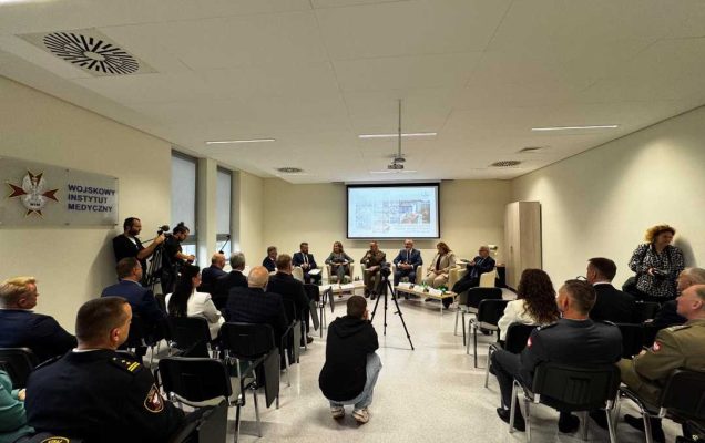 Sala konferencyjna pomiędzy rzędami krzeseł, po środku kucający dziennikarz robi zdjęcie uczestnikom debaty.
