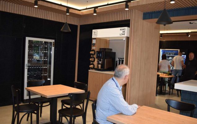 Na pierwszym planie siedzi starszy mężczyzna przy stoliku, za nim widok na dalszą przestrzeń Strefy Smaku. Pomieszczenie ma beżową posadzkę, drewniane ściany oraz głęboko niebieską glazurę na części ścian.