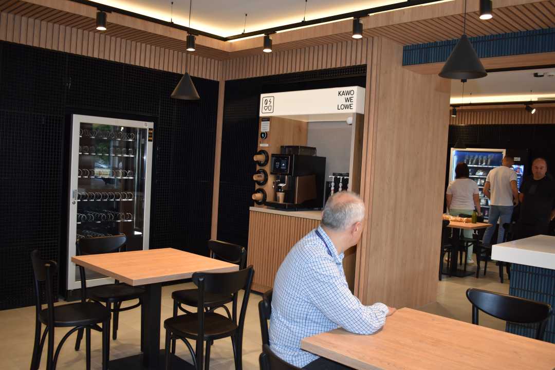 Na pierwszym planie siedzi starszy mężczyzna przy stoliku, za nim widok na dalszą przestrzeń Strefy Smaku. Pomieszczenie ma beżową posadzkę, drewniane ściany oraz głęboko niebieską glazurę na części ścian.