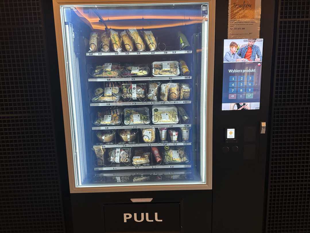 Automat do sprzedaży gotowych posiłków. Maszyna posiada duże szklane drzwi. Przez szybę widać na półkach zapakowane posiłki. Prawa część automatu zawiera wyświetlacz do składania zamówień oraz moduł do płatności.