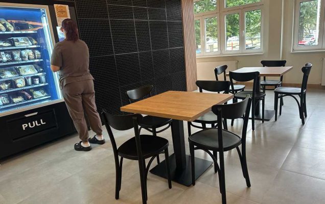 Na pierwszym planie młoda kobieta przed automatem sprzedającym gotowe posiłki , za nią 2 kwadratowe stoliki z krzesłami. Pomieszczenie ma beżową posadzkę, drewniane ściany oraz głęboko niebieską glazurę na części ścian.