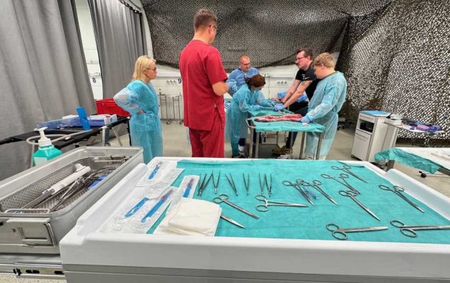 Na pierwszym planie narzędzia medyczne przygotowane do operacji. Na drugim planie grupa lekarzy w sali treningowej nad stołem operacyjnym.