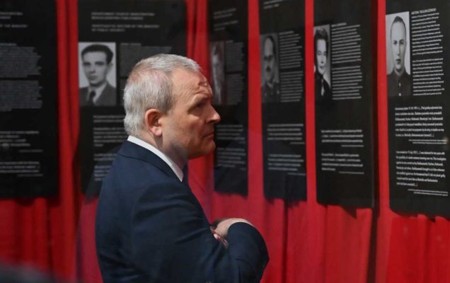 Pracownik WIM ogląda ekspozycję złożoną ze zdjęć i biografii żołnierzy. Plansze mają czarne tło, zawieszone są obok siebie na czerwonych materiałowych kotarach.