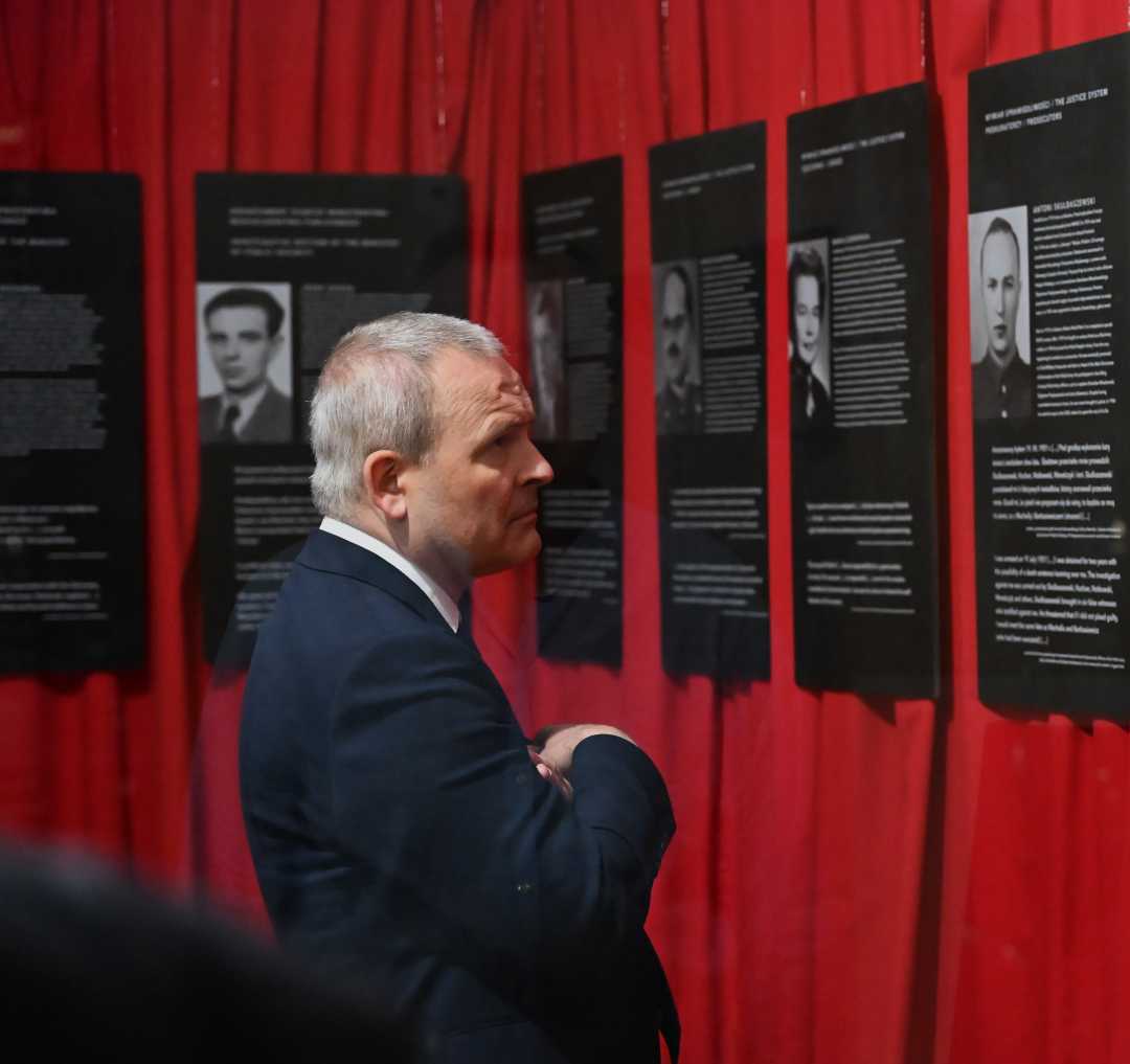 Pracownik WIM ogląda ekspozycję złożoną ze zdjęć i biografii żołnierzy. Plansze mają czarne tło, zawieszone są obok siebie na czerwonych materiałowych kotarach.
