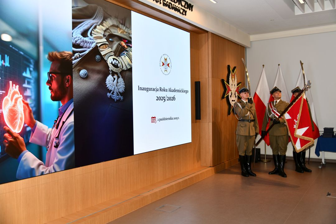 Aula WIM-PIB, trzech żołnierzy w mundurach galowych stoi na baczność z sztandarem Polski, obok nich wyświetlony napis Inauguracja Roku Akademickiego 2025/2026.