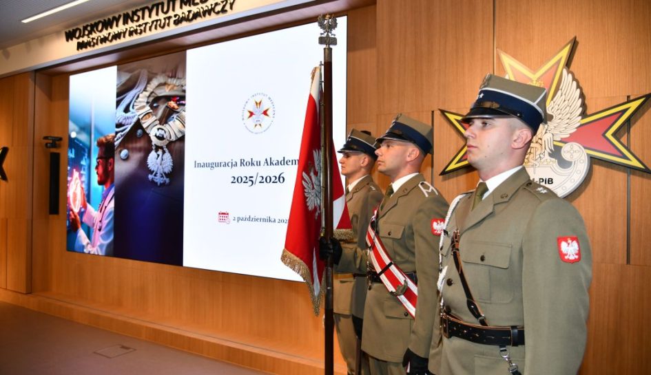 Inauguracja Roku Akademickiego w WIm-PIB. Aula WIM-PIB, trzech żołnierzy w mundurach galowych stoi na baczność z sztandarem Polski, obok nich wyświetlony napis Inauguracja Roku Akademickiego 2025/2026.